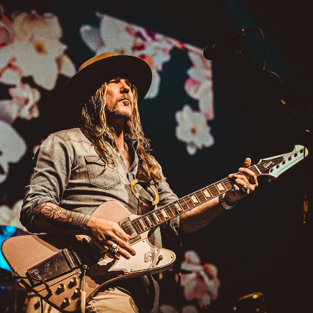 Devon Allman plays the guitar in front of a floral backdrop