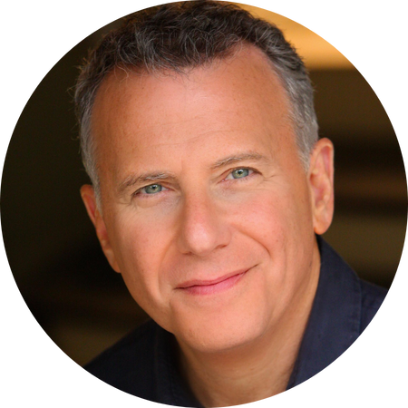 Paul Reiser stares into the camera, a wry grin twisting around his friendly face.  He wears a blue button-down shirt.