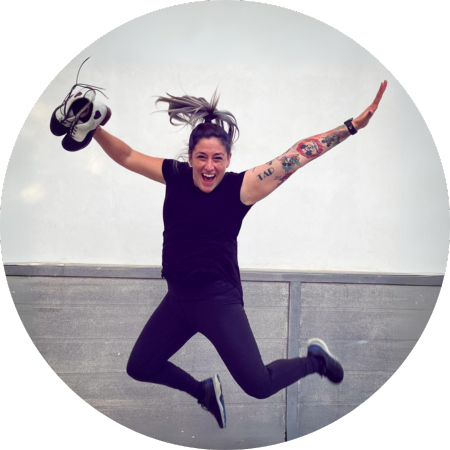 Anita Boyer is captured mid-air in a joyful jump, with one arm stretched high and the other holding a pair of sneakers. They have a broad smile, sporting a black t-shirt and black pants. Their hair is swept back by the motion, and they have visible tattoos on their arm. The background appears to be a plain, light-colored wall, emphasizing the person's dynamic pose.