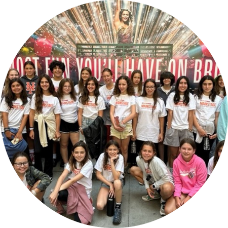 A group of smiling youths gathered for a photo, wearing coordinated t-shirts, in front of a vibrant Broadway show advertising backdrop.