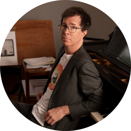 Ben Folds leans with his elbows on the keys of his piano in a blazer and t-shirt.