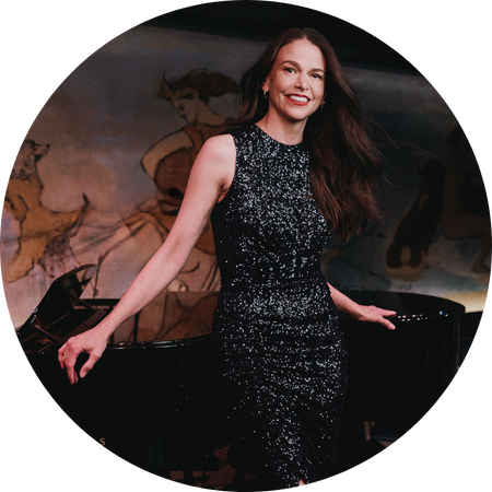 Sutton Foster, a woman with long, flowing brown hair and a radiant smile, poses elegantly in front of a grand piano. She wears a form-fitting, sleeveless black sequined dress that shimmers under the soft lighting. The background features an artistic mural with classical figures, adding a timeless and sophisticated ambiance to the scene. Her confident and graceful stance exudes warmth and charm.