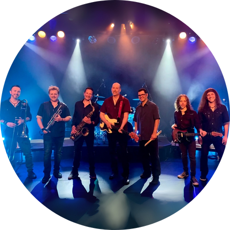 A seven-member band poses on a dimly lit stage with atmospheric lighting casting beams of white and blue behind them. The musicians, dressed in dark attire, hold various instruments, including a trombone, trumpet, saxophone, electric guitar, bass, and drumsticks. The central figure, a bald man wearing a red shirt, holds an electric guitar and stands confidently in the spotlight. The group exudes a strong stage presence, ready to perform.