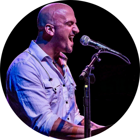 Mike DelGuidice passionately sings into a microphone while playing the piano on stage. He wears a white button-up shirt with the sleeves rolled up, revealing tattoos on his arms. His facial expression conveys deep emotion, and the stage lighting casts a dramatic blue and purple glow on him, enhancing the intensity of the performance.