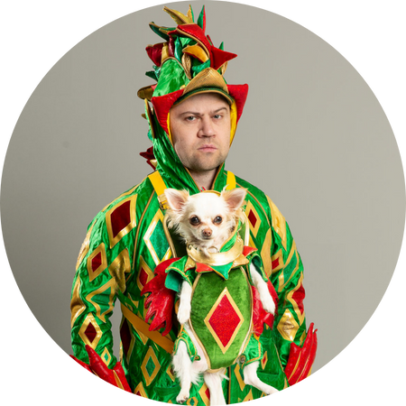 A man dressed in a vibrant, green and red dragon costume with gold diamond patterns stands against a plain gray background, wearing a serious expression. A small Chihuahua, also dressed in a matching dragon outfit, is secured in a front-facing pet carrier, looking directly at the camera. The elaborate and whimsical costumes contrast humorously with the man's deadpan expression.