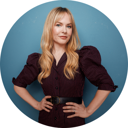 Rosebud Baker, a confident woman with long, wavy blonde hair, poses against a textured blue background. She wears a stylish, dark pinstriped dress with puffed sleeves and a cinched waist, accentuated by a black belt with a circular buckle. Her hands rest on her hips, and she has a poised expression with a slight smile, exuding elegance and self-assurance.