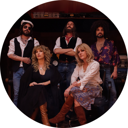 A group of five musicians pose in a recording studio, dressed in 1970s-inspired bohemian rock attire. The two women sit in the foreground, one wearing a black flowing outfit with curly blonde hair, and the other in a white blouse, patterned skirt, and knee-high suede boots. The three men stand behind them, sporting vests, patterned shirts, and accessories like a captain’s hat and sunglasses. The studio setting, with mixing equipment in the background, enhances the vintage rock aesthetic.