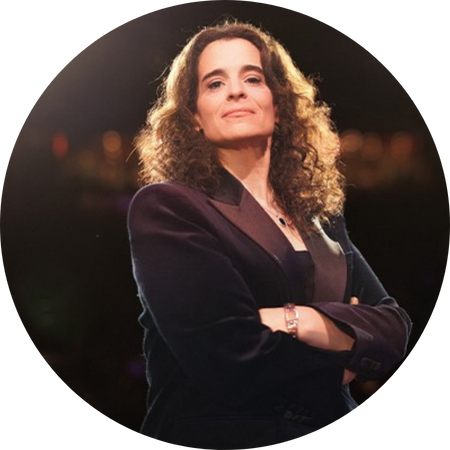 Jessica Kirson, a confident woman with shoulder-length curly hair, stands with her arms crossed, exuding authority and poise. She wears a dark, tailored blazer over a black top, accessorized with a silver bracelet and necklace. The warm stage lighting highlights her face, creating a dramatic effect against the softly blurred background.