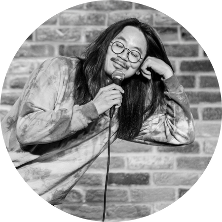 A black-and-white photograph of Sheng Wang, a comedian with long hair and round glasses, performing on stage. He wears a tie-dye sweatshirt and leans playfully on an invisible surface while holding a microphone, with a relaxed and amused expression. The brick wall background suggests a classic comedy club setting, enhancing the intimate and laid-back atmosphere.