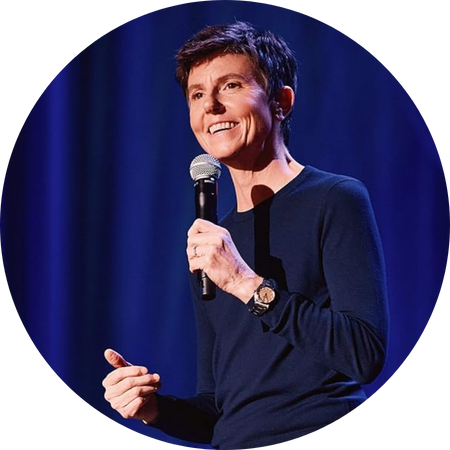 A comedian performs on stage, holding a microphone and smiling warmly. She wears a dark, long-sleeved shirt and a wristwatch, with short brown hair neatly styled. The stage is lit with a deep blue curtain backdrop, creating a professional yet intimate setting. Her relaxed posture and expressive gestures convey a natural and engaging presence.