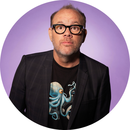 Tom Papa, a middle-aged man with short, slightly receding hair and black-framed glasses, poses against a soft purple background. He has a playful, surprised expression with raised eyebrows and wide eyes. He wears a black blazer over a graphic T-shirt featuring a detailed blue octopus illustration. His casual yet stylish look, combined with his expressive face, conveys a fun and charismatic personality.