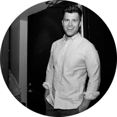 A black-and-white photo of Colin Jost standing casually in a doorway, smiling warmly at the camera. He wears a light-colored, button-up shirt with the sleeves rolled up and has his hands in his pockets. His short, neatly styled hair and confident yet approachable demeanor give the image a candid, behind-the-scenes feel.