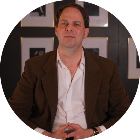 A man in a brown blazer and white dress shirt sits confidently with his hands loosely clasped in front of him. He wears a gold watch and has neatly combed dark hair. Behind him is a black wall adorned with a grid of framed black-and-white photographs, each with a gold border, creating a refined and artistic backdrop. The lighting is warm and focused, suggesting an interview or formal portrait setting.