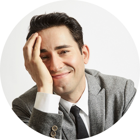 John Lloyd Young, a well-dressed man with dark, neatly styled hair, smiles warmly at the camera. He rests his head on one hand in a relaxed and approachable pose. He wears a gray suit jacket, white shirt, and black tie against a clean white background, giving the image a polished and professional appearance.
