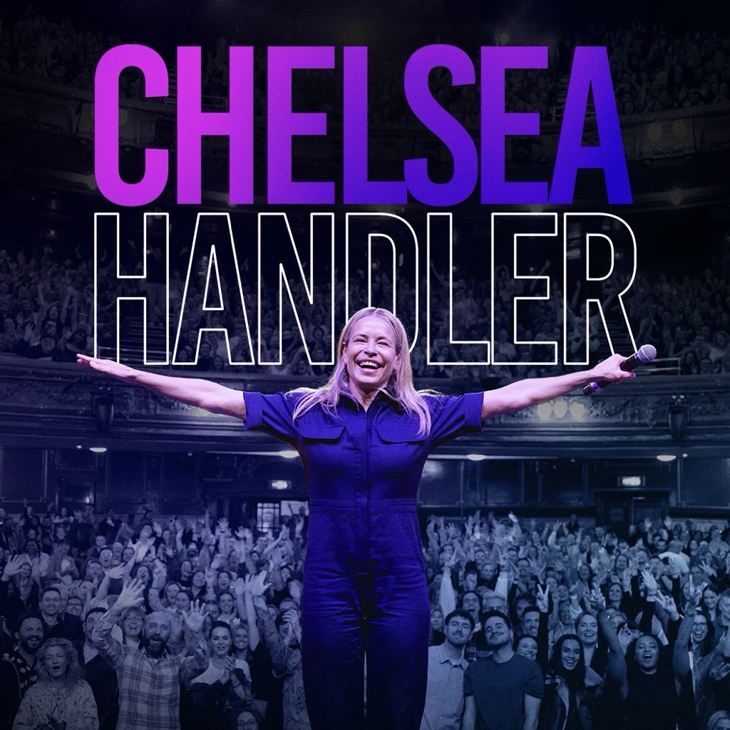 Chelsea Handler, a woman with wavy blonde hair, smiles brightly at the camera, wearing a cozy dark colored sweater. She rests her chin gently on her hand in a relaxed and friendly pose, with soft lighting and a plain light gray background enhancing the warm, approachable feel of the portrait.