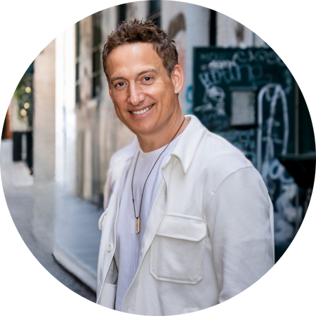 Elon Gold, a man with short, brown hair and a warm smile poses for a headshot. He is wearing a white t-shirt and a white button-up shirt over it, with a necklace that has a small, rectangular pendant. The background is a slightly blurred outdoor scene with what appears to be an alleyway and some graffiti on a wall.