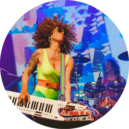 A circular action shot of the 80s cover band Jessie's Girl performing live. In the foreground, a female musician with voluminous curly hair, sunglasses, and neon green attire plays a white keytar with a rainbow strap. In the background, a drummer is visible behind a drum kit amidst vibrant blue and purple stage lighting.
