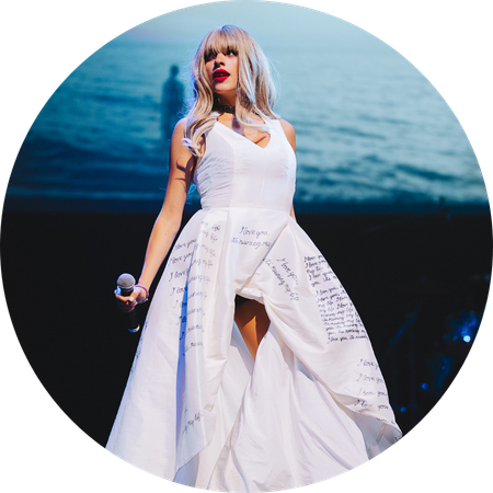 A performer from the tribute act “Let’s Sing Taylor” stands on stage holding a microphone, wearing a flowing white dress with handwritten-style text on the skirt, with a large ocean scene projected in the background.