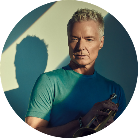 Chris Botti, a man with short blond hair wearing a teal T-shirt, holds a trumpet while standing in dramatic light and shadow against a plain wall.